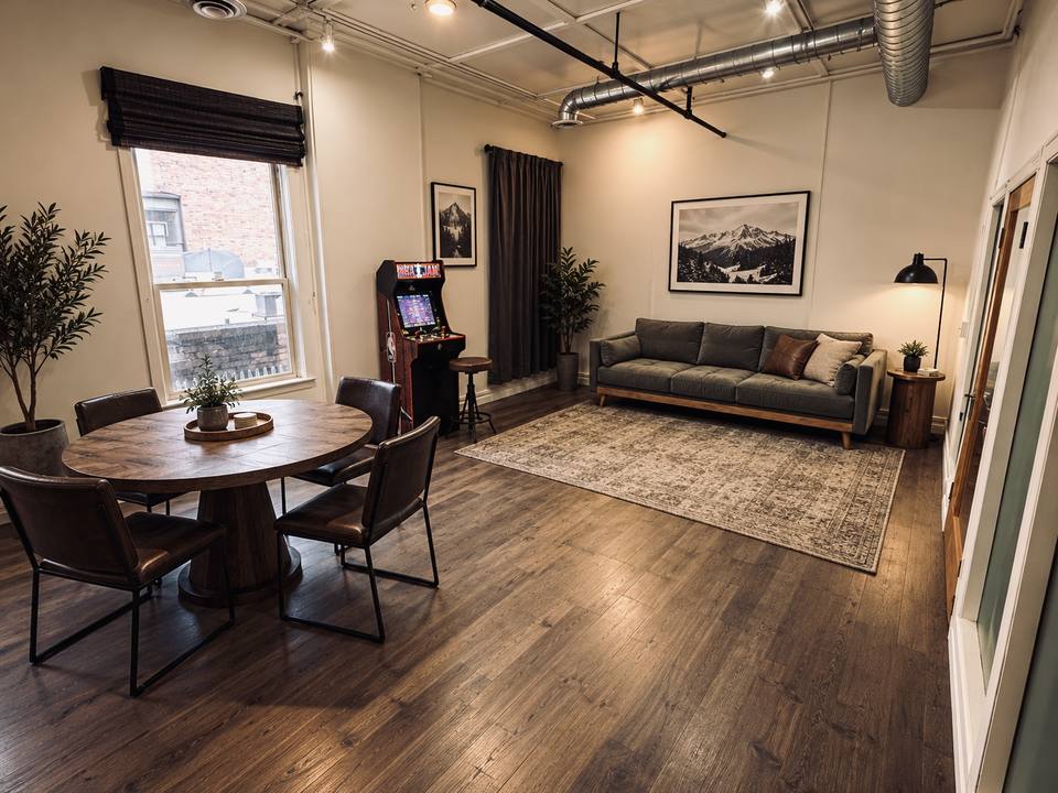 The Parlor set with a round wood table and leather chairs, a grey sofa on a rug, an arcade cabinet and a framed mountain print beside tall windows.