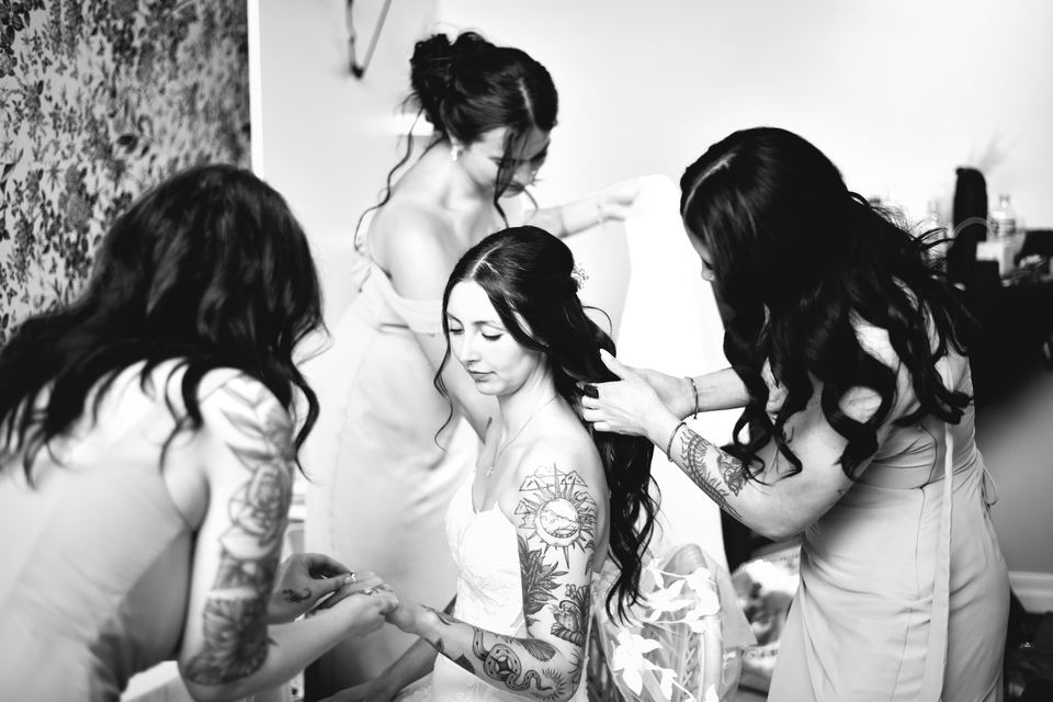 A black-and-white photo of a bride sitting while her bridesmaids help her get ready, one fixing her hair and another beside her.