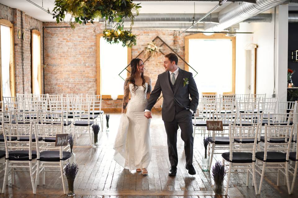 A couple in wedding clothes walking hand in hand up the aisle of the Loft, between rows of white chairs, with a diamond arch and tall windows behind them.