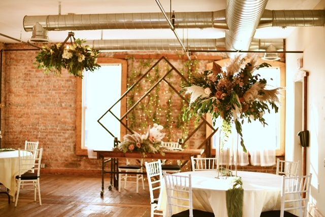 A sweetheart table under a tall arrangement of pampas grass and roses in the Loft, with round tables and a greenery chandelier.