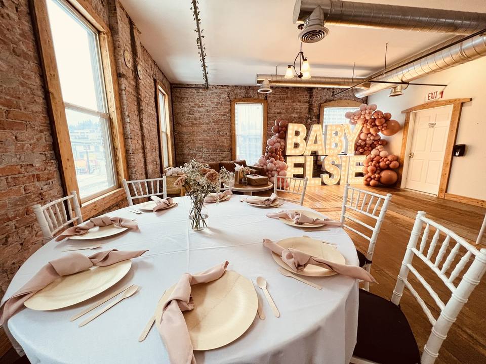 A baby shower in the Loft: round tables with wooden plates and blush napkins, and a lit BABY sign with balloons on the brick wall.