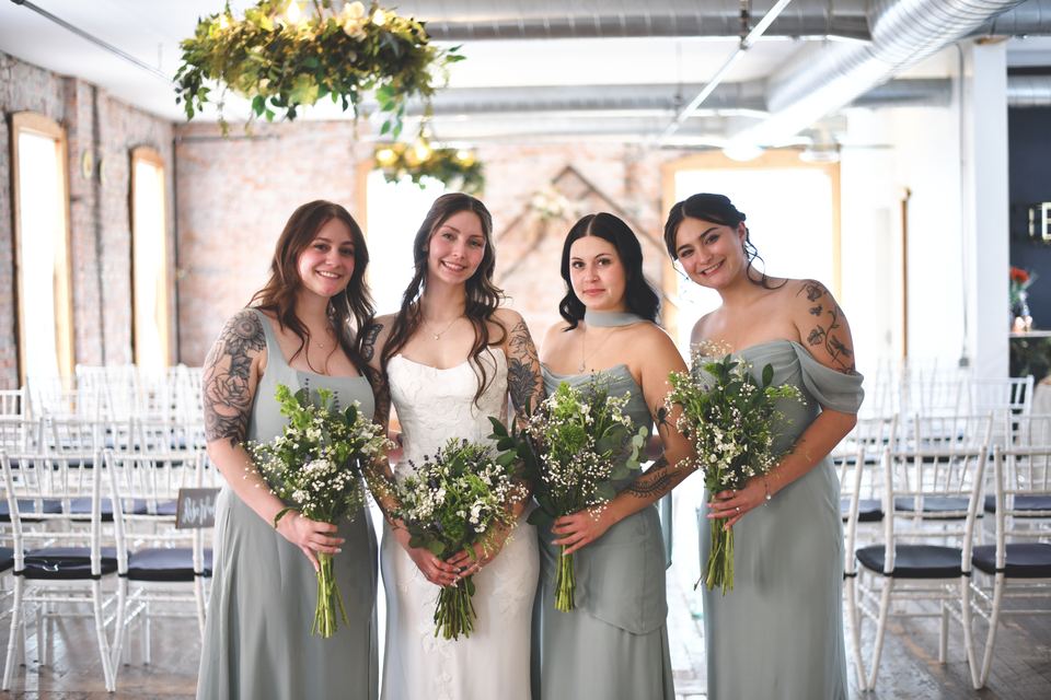 A bride in white standing with three bridesmaids in sage green holding bouquets of white flowers, with rows of chairs and a greenery chandelier in the Loft behind them.