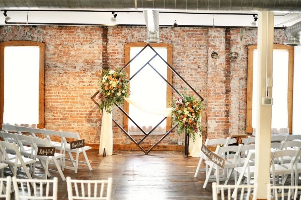 A diamond-shaped arch with flowers and draped fabric between two Loft windows, with rows of white chairs marked Bride and Groom.