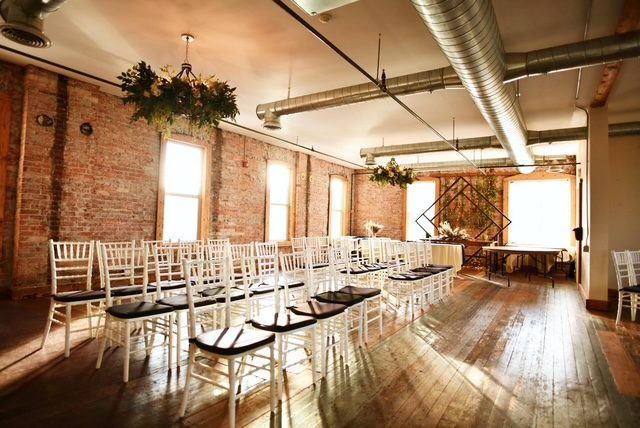 The Loft set for a ceremony: rows of white chiavari chairs facing a diamond arch between the windows, with a greenery chandelier overhead.