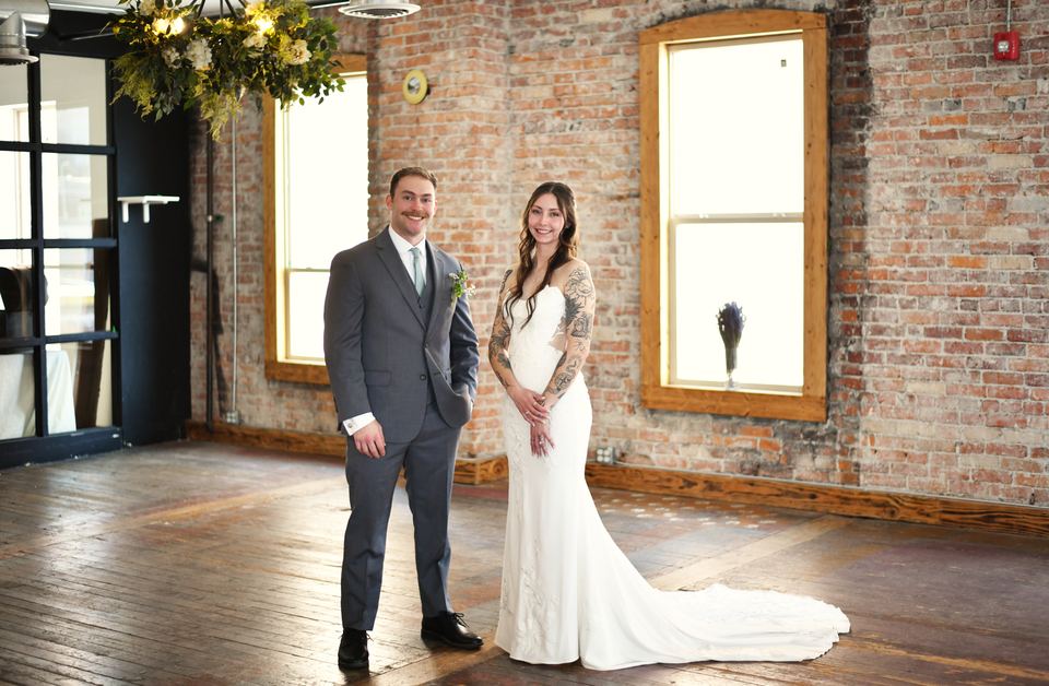 A couple in wedding clothes standing side by side in the Loft, in front of a brick wall with tall windows.