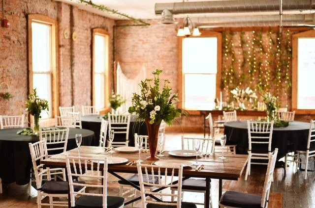 A wooden farm table with tall greenery beside round tables in black cloths in the Loft.