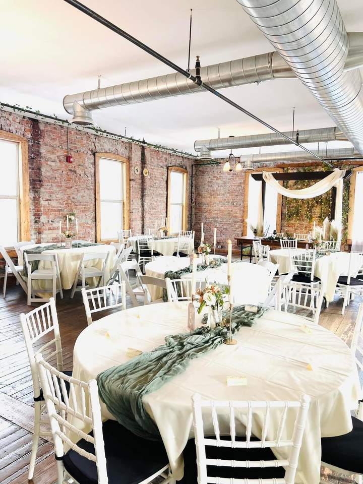 Round tables in the Loft with cream cloths, sage runners, taper candles and white chiavari chairs.