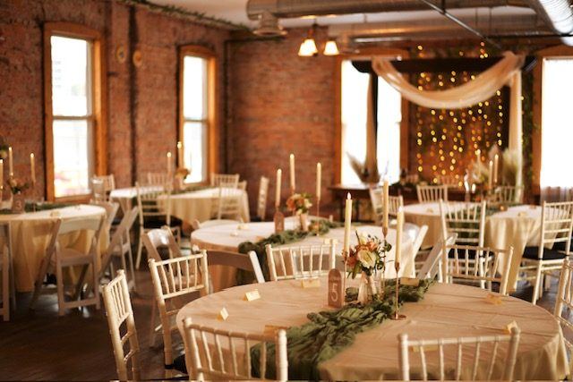 The Loft set for a reception: round tables with candles and greenery runners, and a string-light backdrop draped in white.