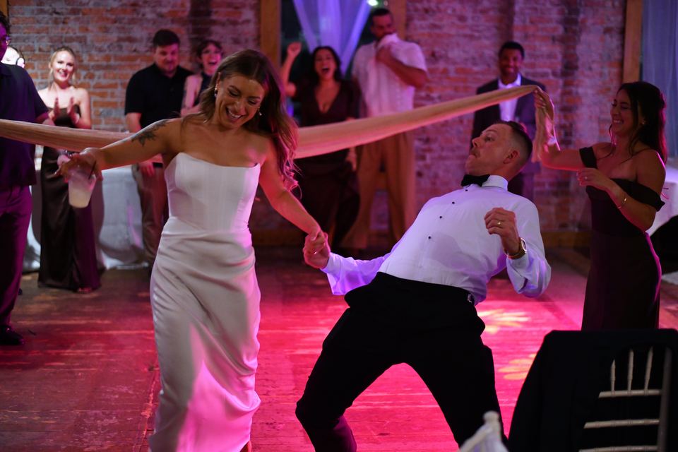 A woman in a white dress and a man in a bow tie dancing on a wood floor under pink light, with a length of fabric stretched above them and guests watching in front of the brick wall.
