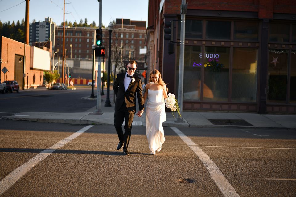 A couple in wedding clothes holding hands and crossing a downtown Spokane street at sunset, with brick buildings behind them.